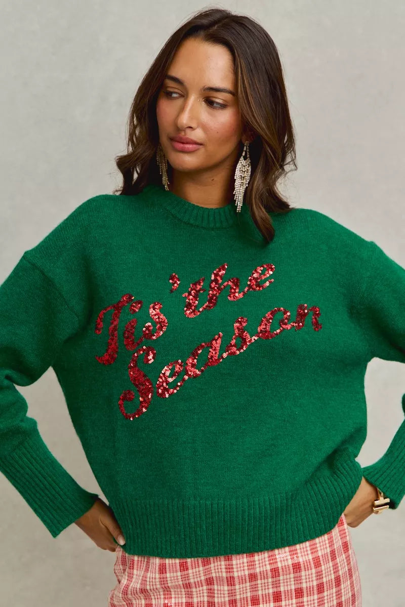 Woman wearing a green sweater with 'Tis the Season' text in red sequins against a neutral background