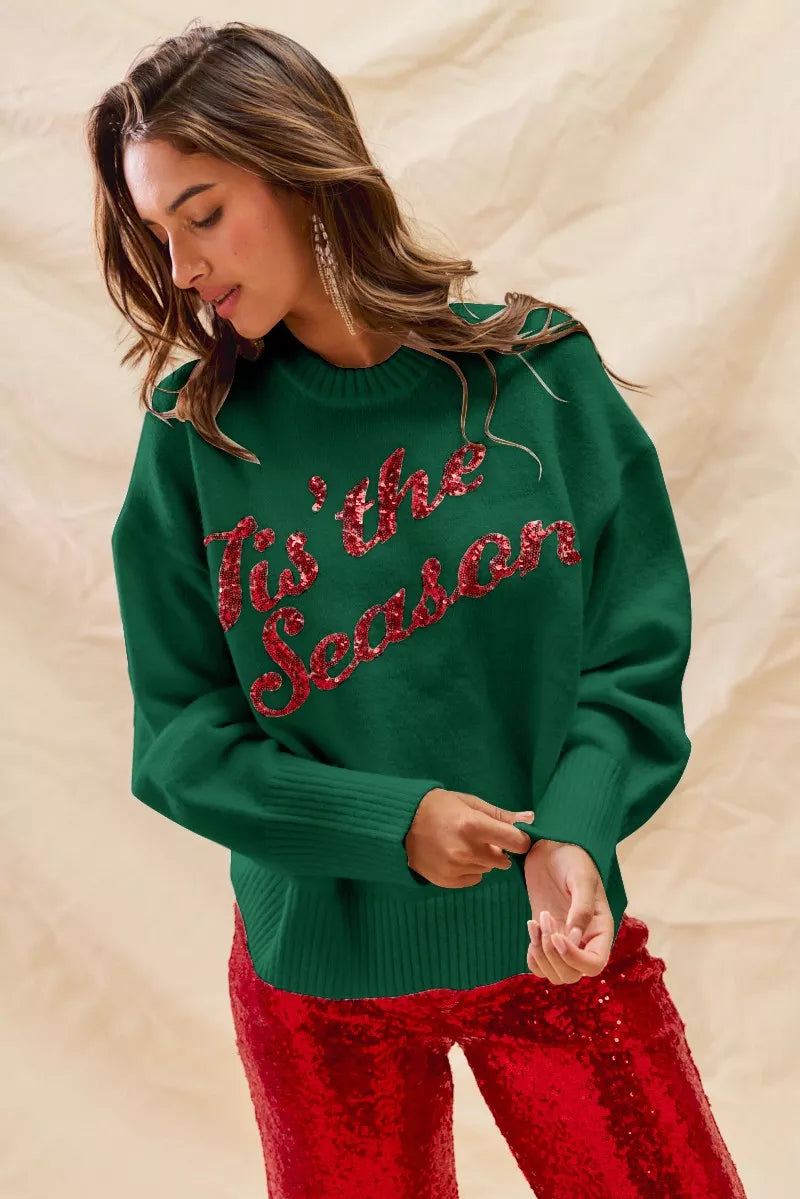 Woman wearing a green sweater with red sequins saying tis the season and red pants against a beige background