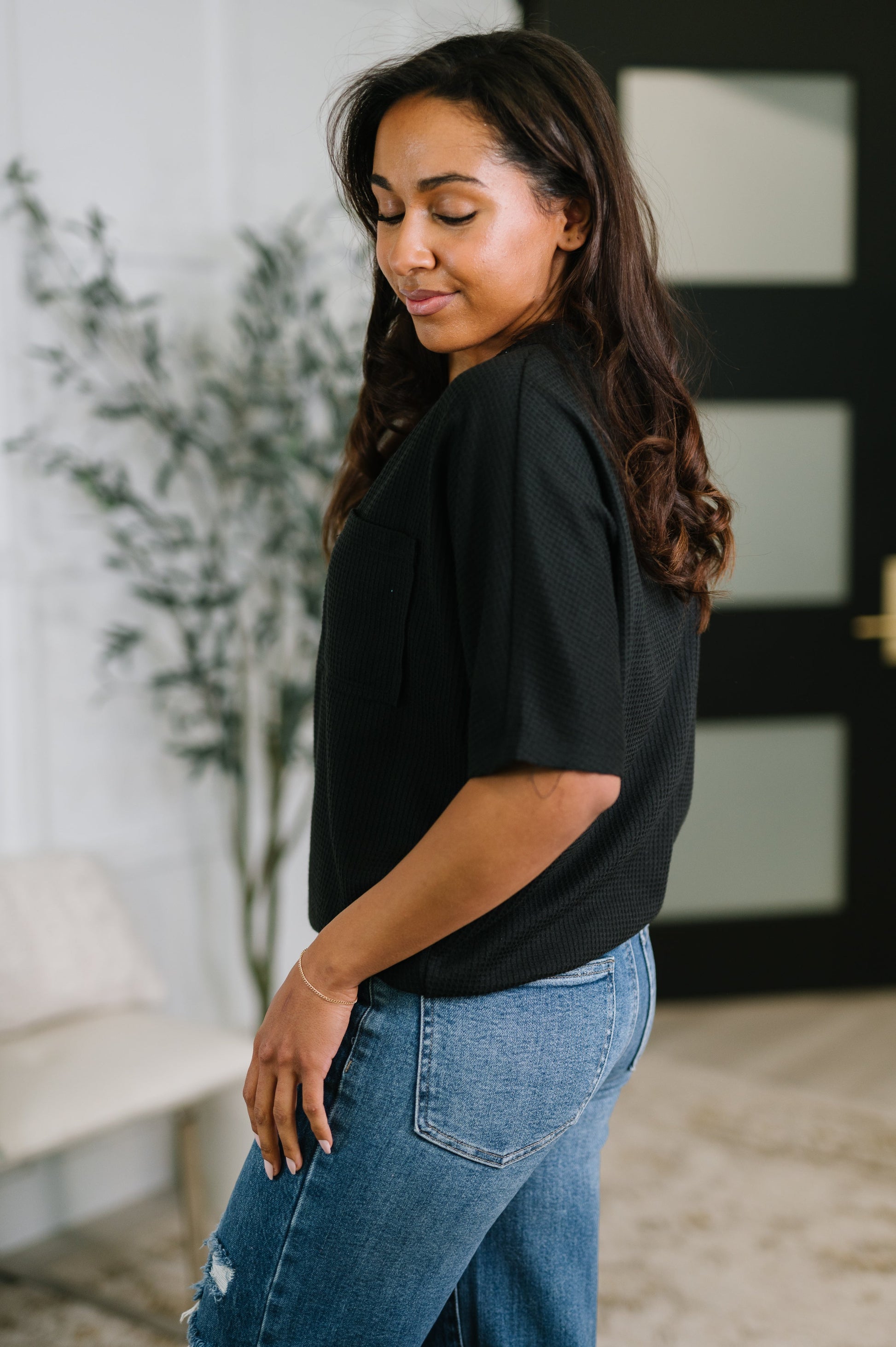 Woman wearing a black top and blue jeans standing indoors with a plant and wall decor in the background.