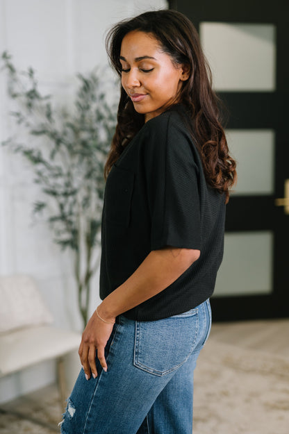 Woman wearing a black top and blue jeans standing indoors with a plant and wall decor in the background.