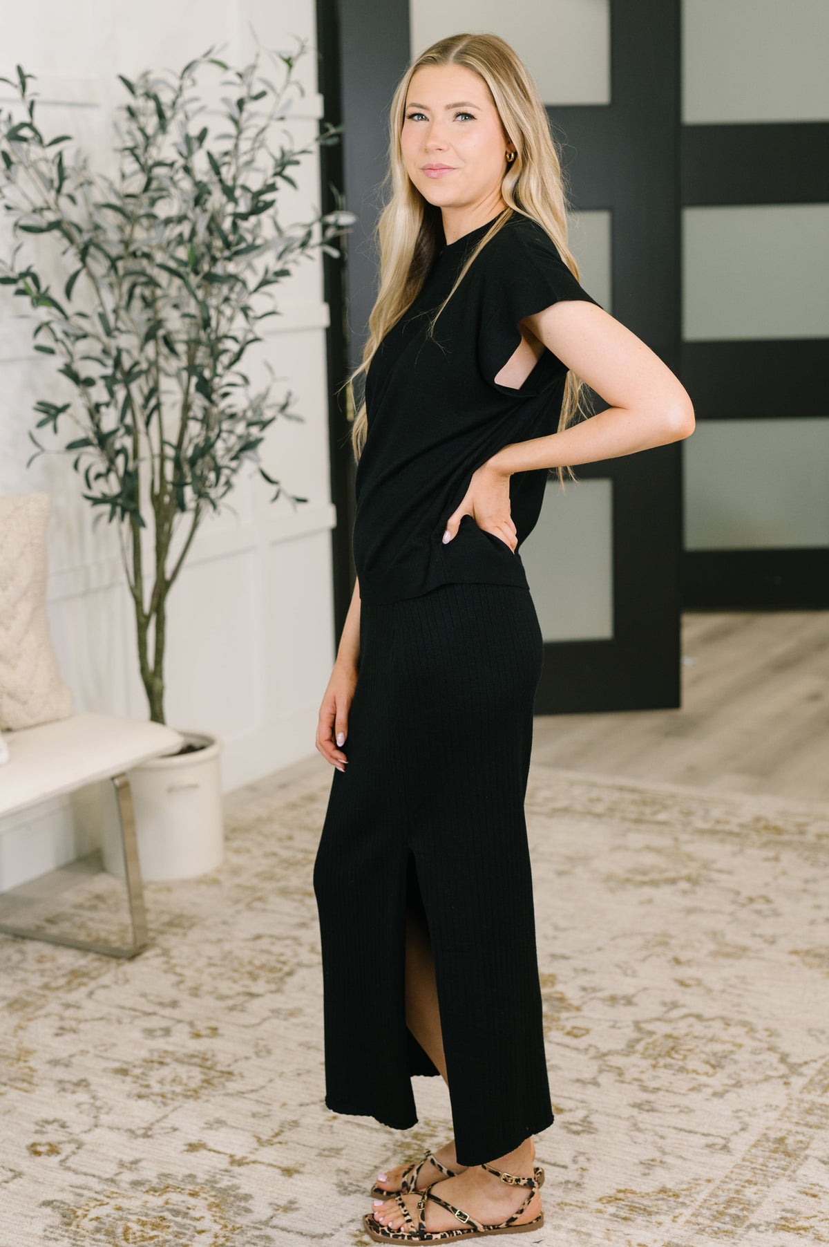 A woman modeling a black short-sleeve maxi dress with side cutouts and a front slit, standing in a modern indoor setting with a patterned rug, potted plant, and black-and-white paneled door.