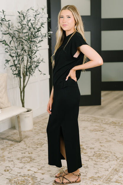 A woman modeling a black short-sleeve maxi dress with side cutouts and a front slit, standing in a modern indoor setting with a patterned rug, potted plant, and black-and-white paneled door.