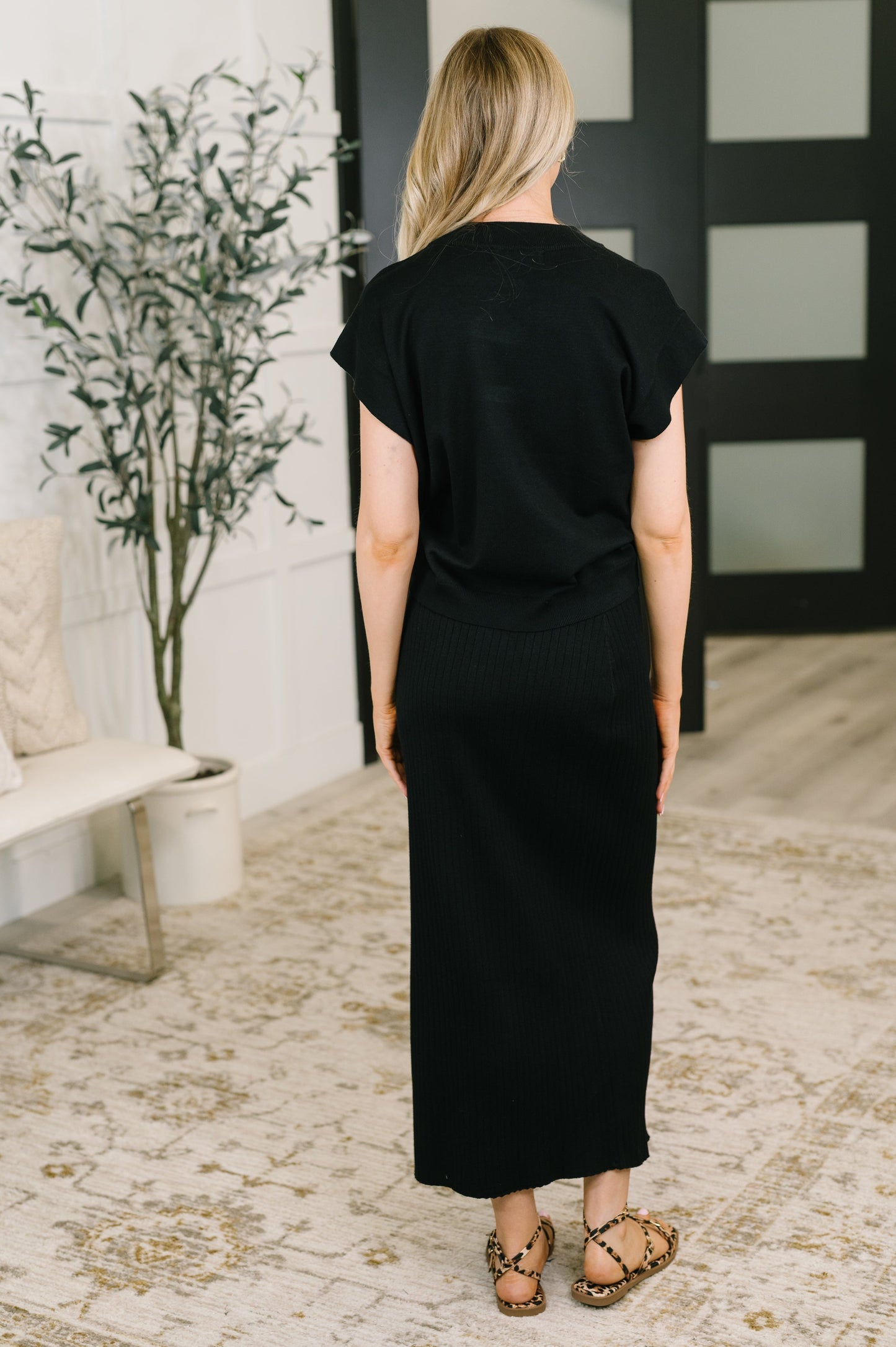 A woman viewed from behind wearing a black short-sleeve maxi dress in a bright, modern room with a patterned rug and a potted plant.