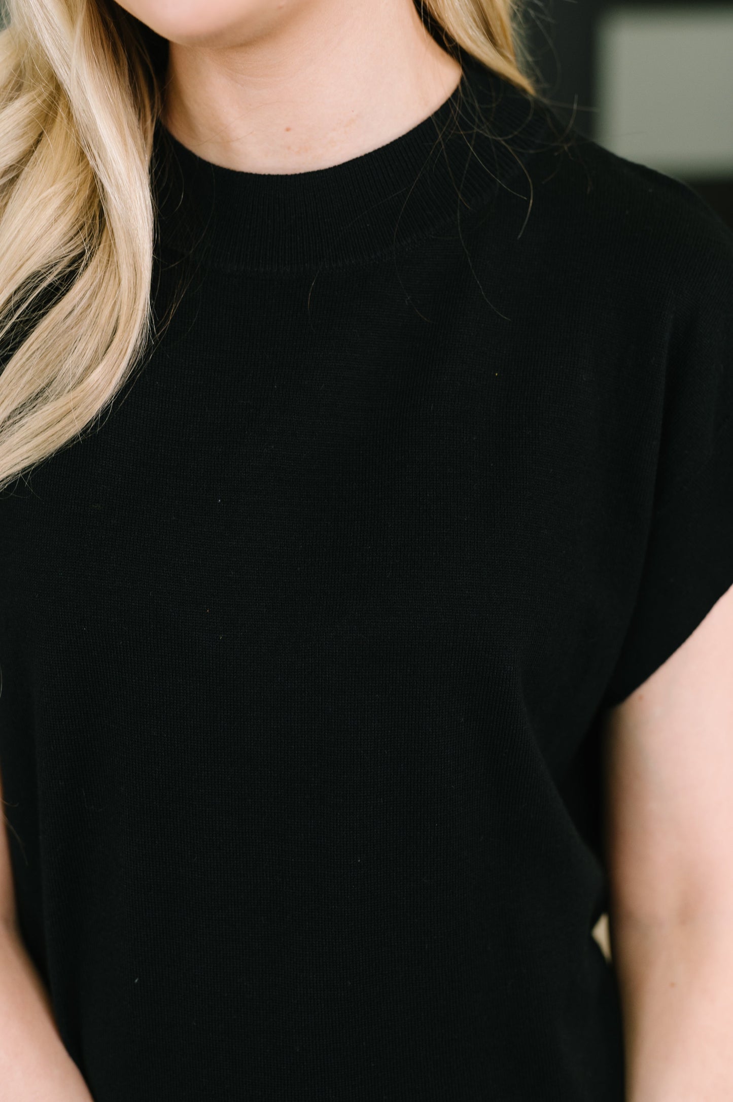 Close-up of a person wearing a plain black short-sleeve crew-neck top, with blonde hair visible at the left edge.