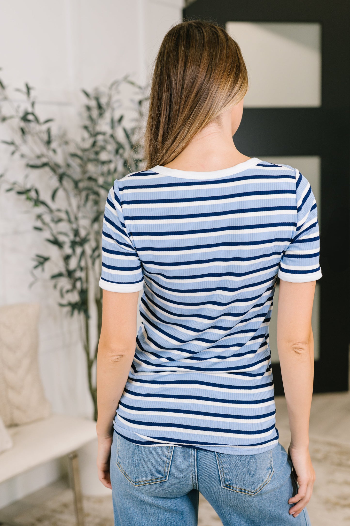 A woman viewed from behind wearing a short-sleeve crewneck t-shirt with horizontal navy and light blue stripes, paired with light wash blue jeans in a bright indoor setting.