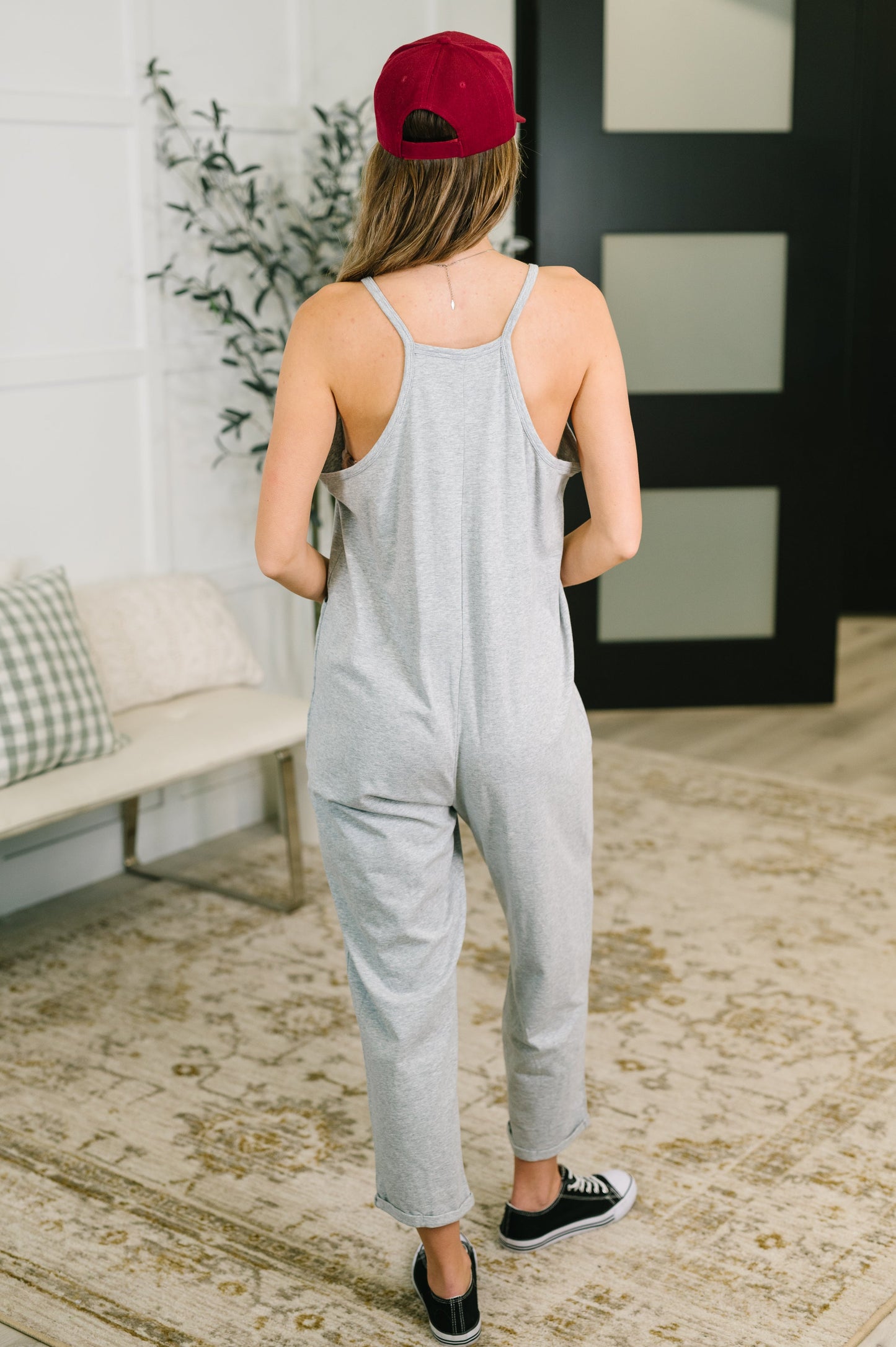 Person wearing a gray jumpsuit and red cap in a room with a couch and plant.