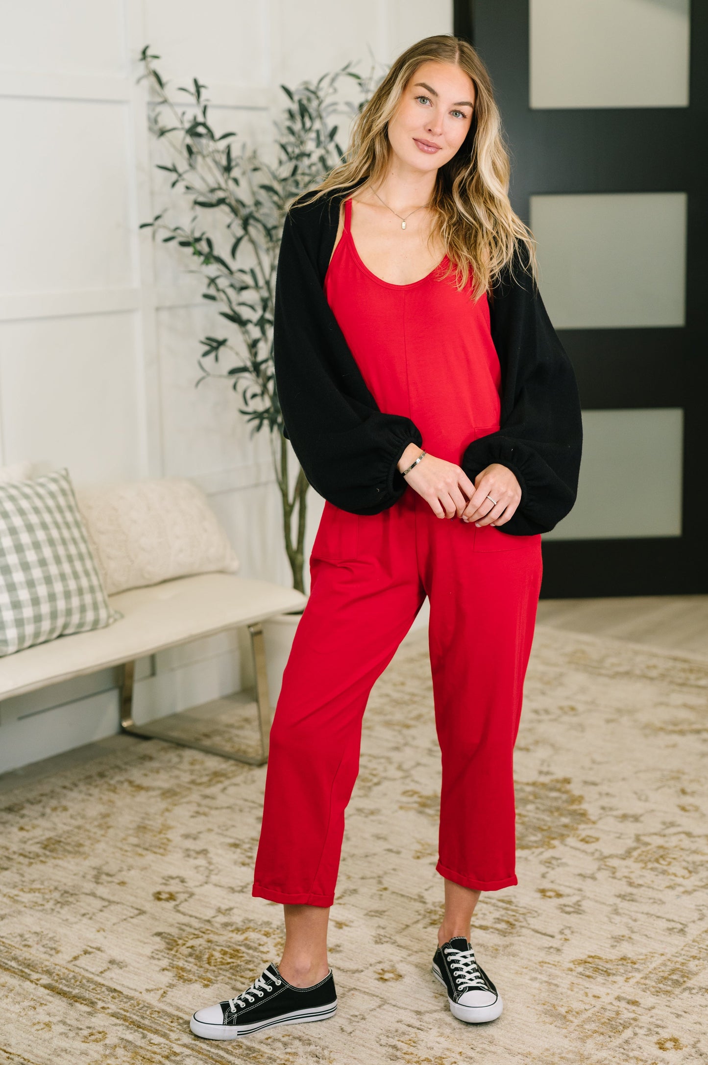Woman wearing a black cardigan and red pants standing in a room with a plant and bench.