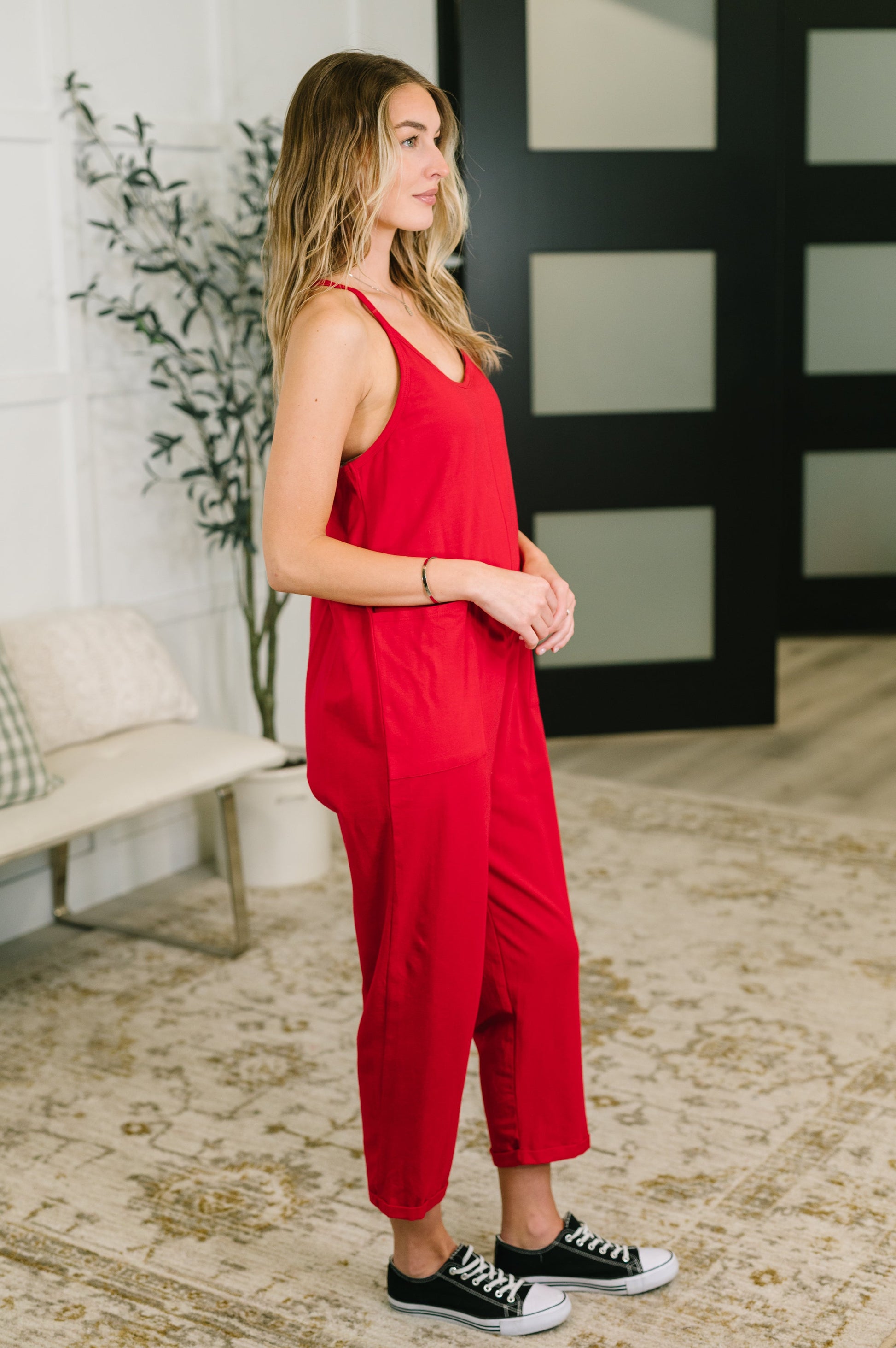 Woman wearing a red jumpsuit standing in a room with a plant and bench.