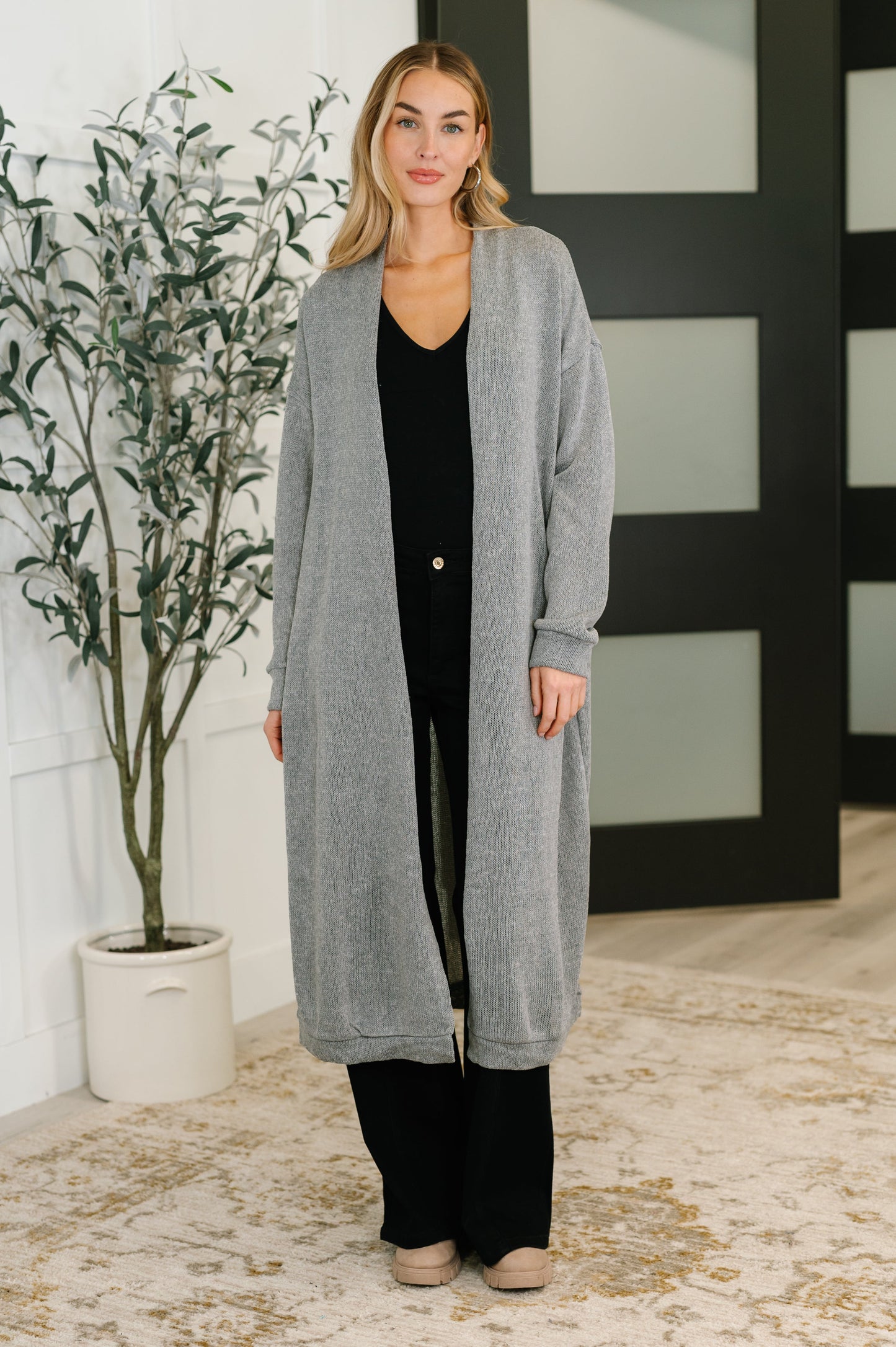 Woman wearing a long gray cardigan in an indoor setting with a plant and wall decor.