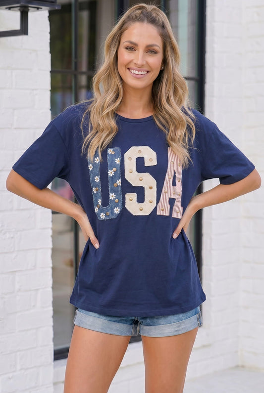 Woman wearing a navy blue t-shirt with 'USA' printed on it, standing against a white brick wall.