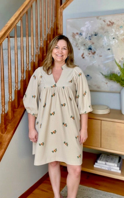 Woman in a beige dress with floral patterns standing in a home setting.