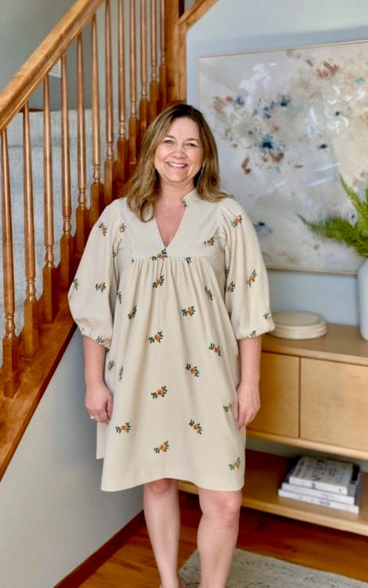Woman in a beige dress with floral patterns standing in a home setting.