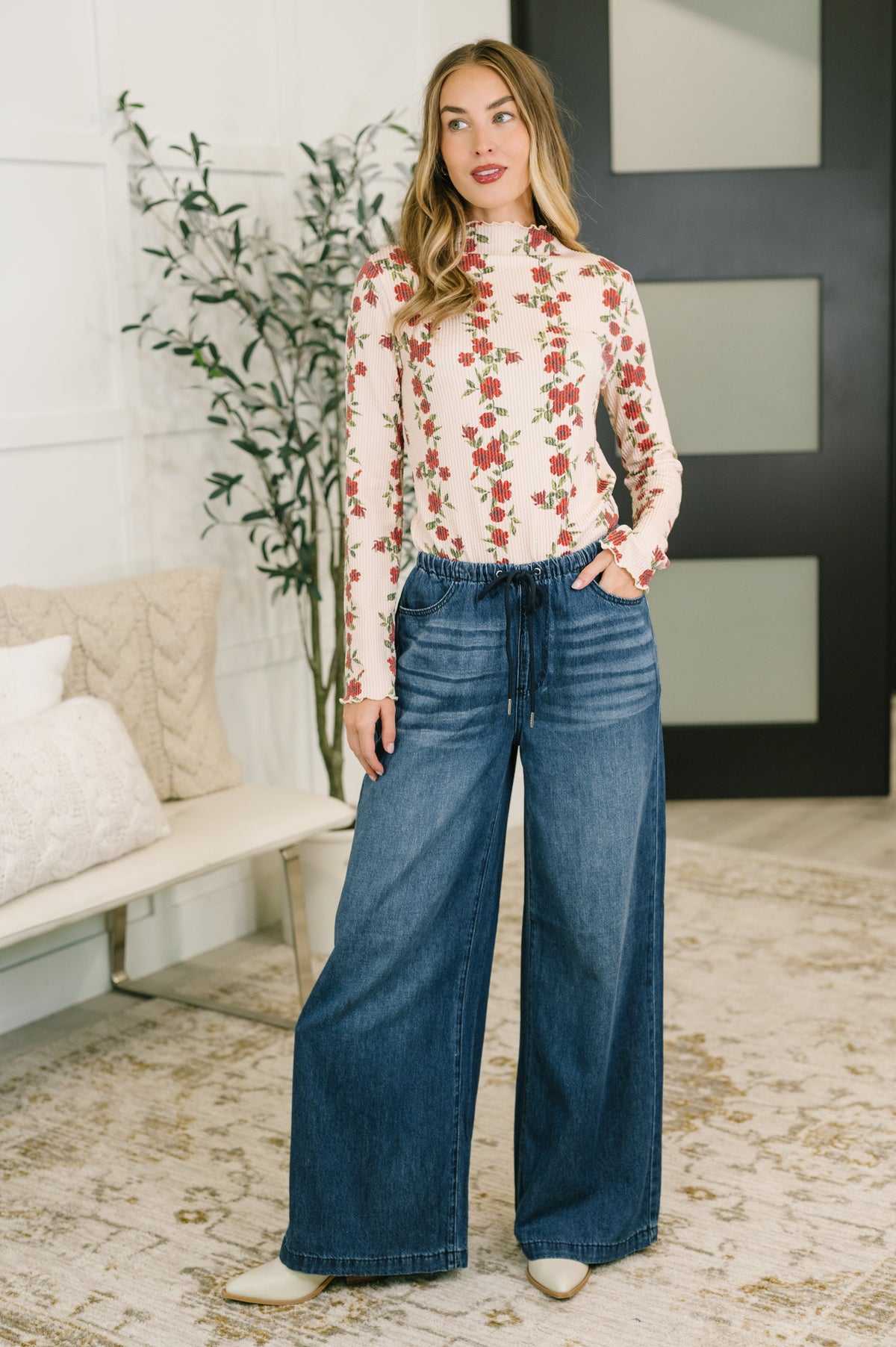 A woman stands in a bright indoor room wearing a cream long-sleeve mock-neck blouse with a red and green floral print, paired with high-waisted wide-leg blue jeans and white pointed-toe boots.