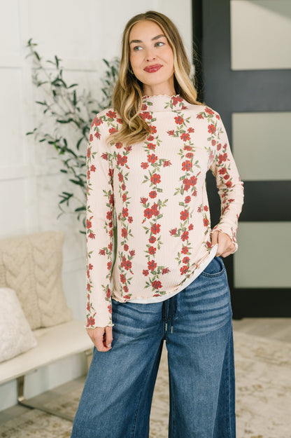 A woman wearing a long-sleeve mock neck top with a red rose floral pattern on a cream background, paired with blue jeans, standing in a brightly lit indoor setting.