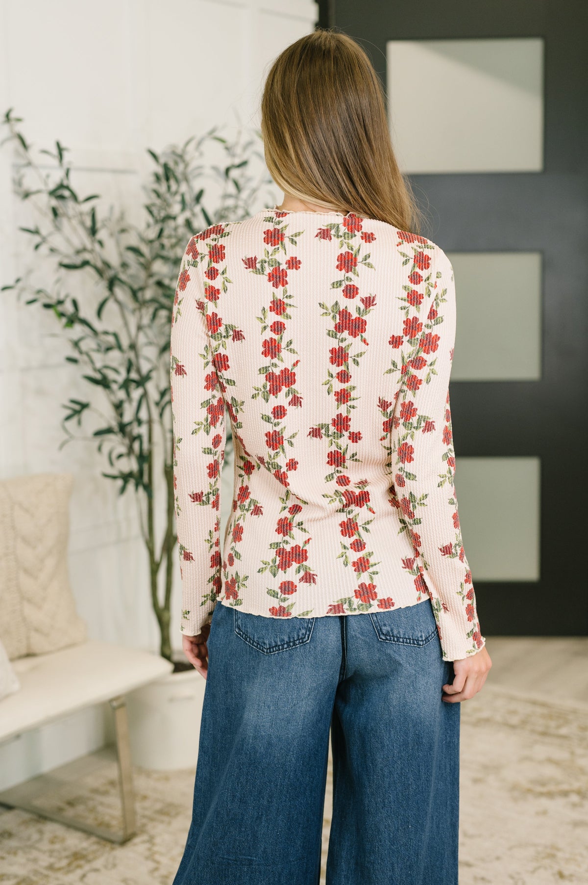 A woman viewed from behind wearing a long-sleeve cream-colored top with a red and green floral vine pattern, paired with blue wide-leg jeans in a modern indoor setting.