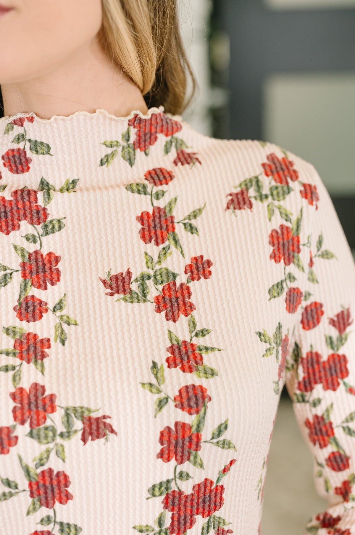 Close-up of a person wearing a ribbed long-sleeve top with a cream base and an allover red rose and green leaf floral pattern.