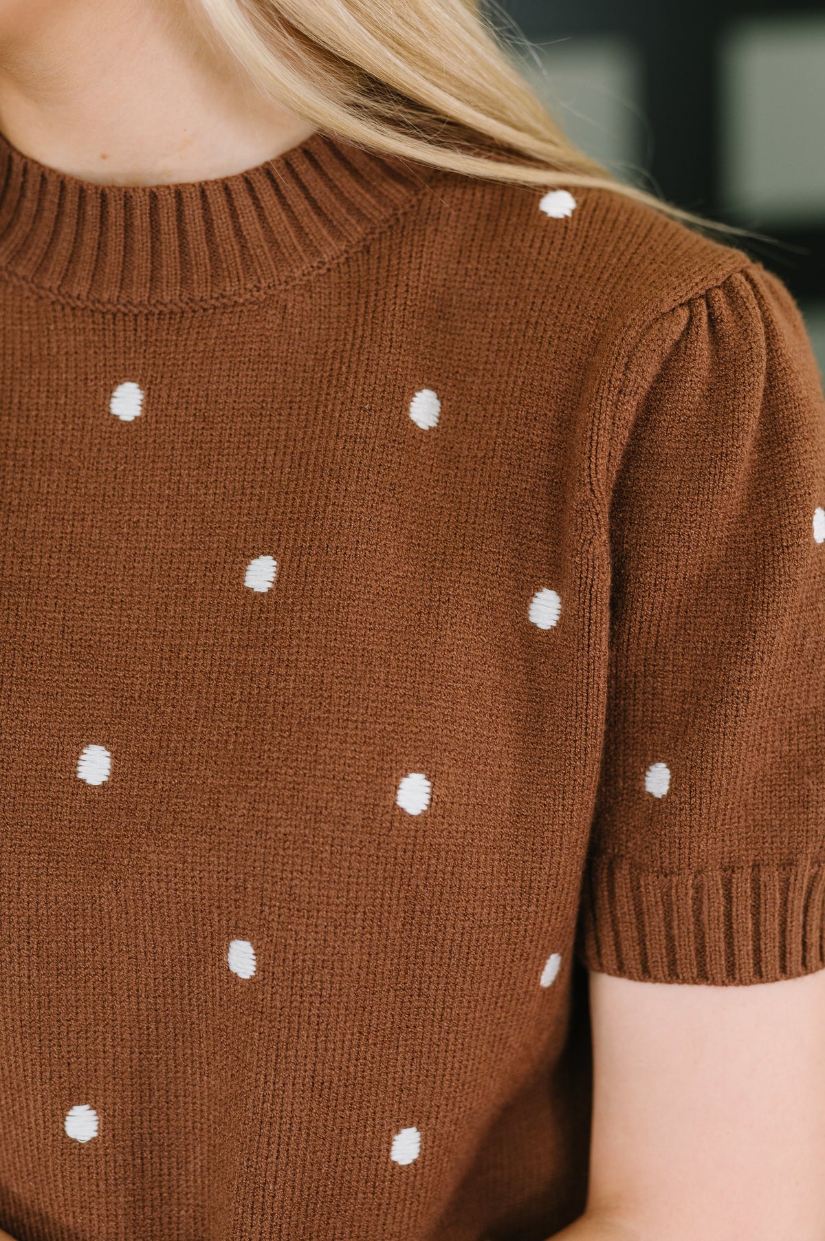 Close-up of a person wearing a brown short-sleeve knit sweater with small white polka dots, showing the chest, shoulder, sleeve, and part of the arm.