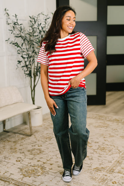 A woman modeling a red and white horizontally striped short-sleeve t-shirt, paired with dark blue jeans and black sneakers, standing in a modern indoor setting with a patterned rug and a potted plant.