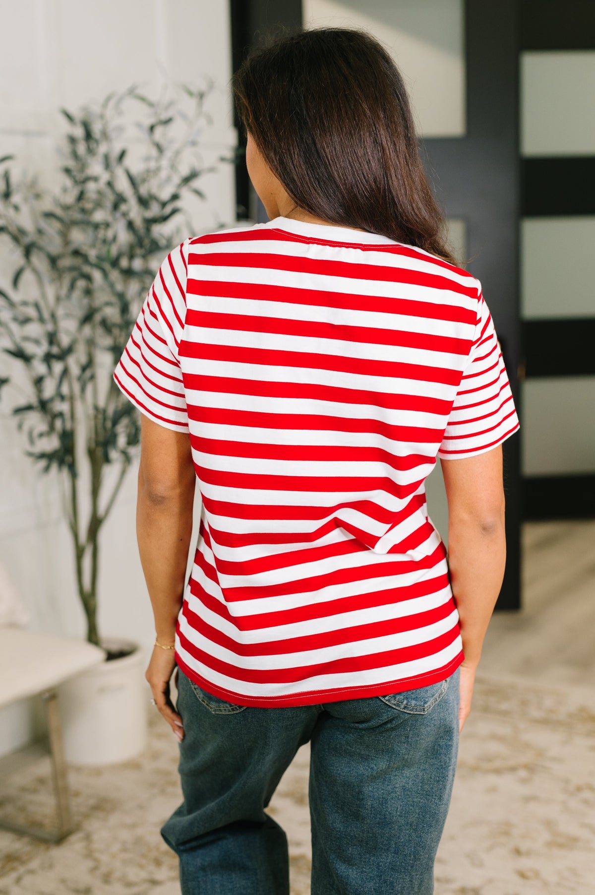 A woman viewed from behind wearing a red and white horizontally striped short-sleeve t-shirt and blue jeans in an indoor setting.