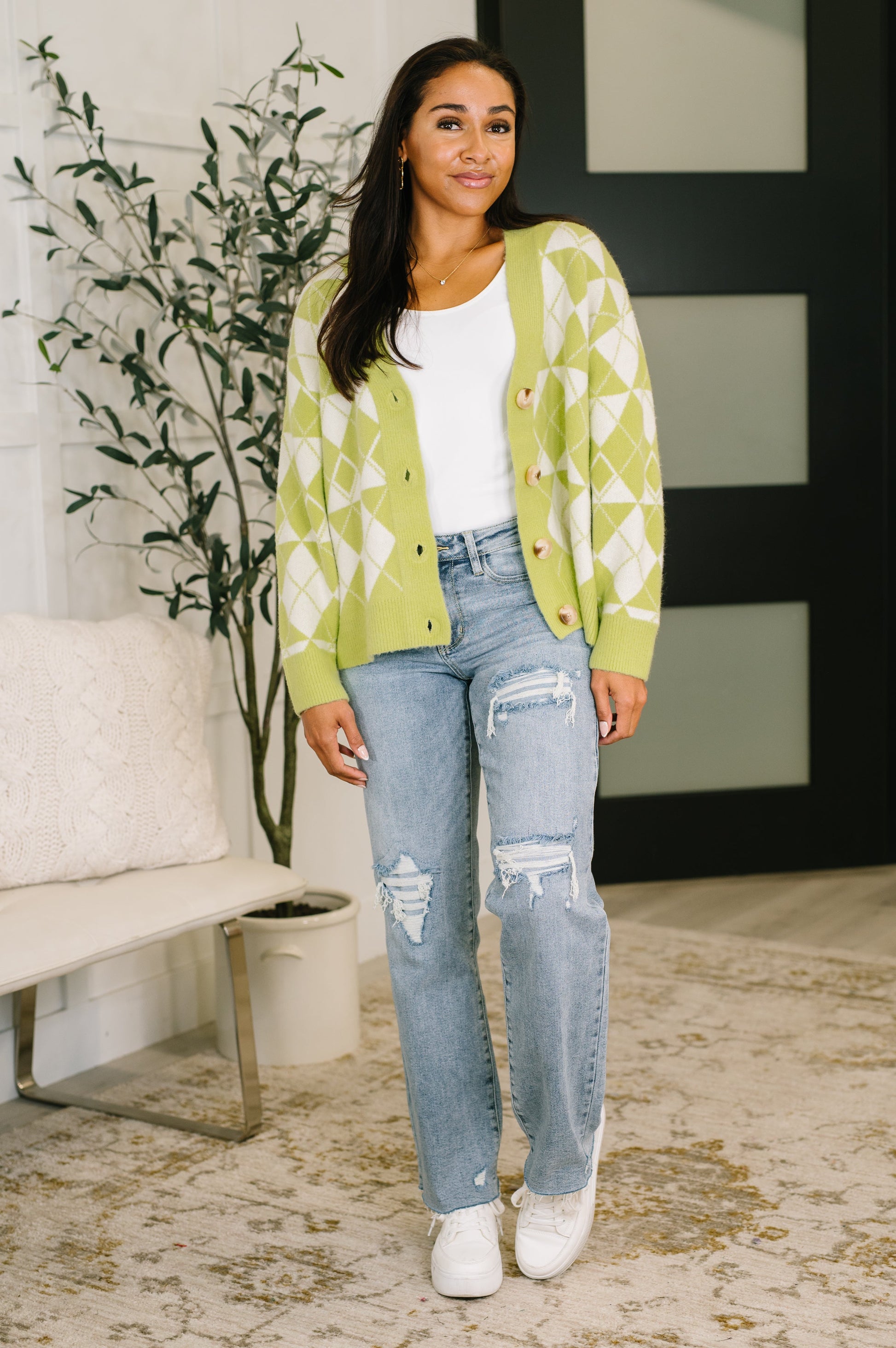 Woman wearing a green patterned cardigan, white shirt, and blue jeans in a living room setting.
