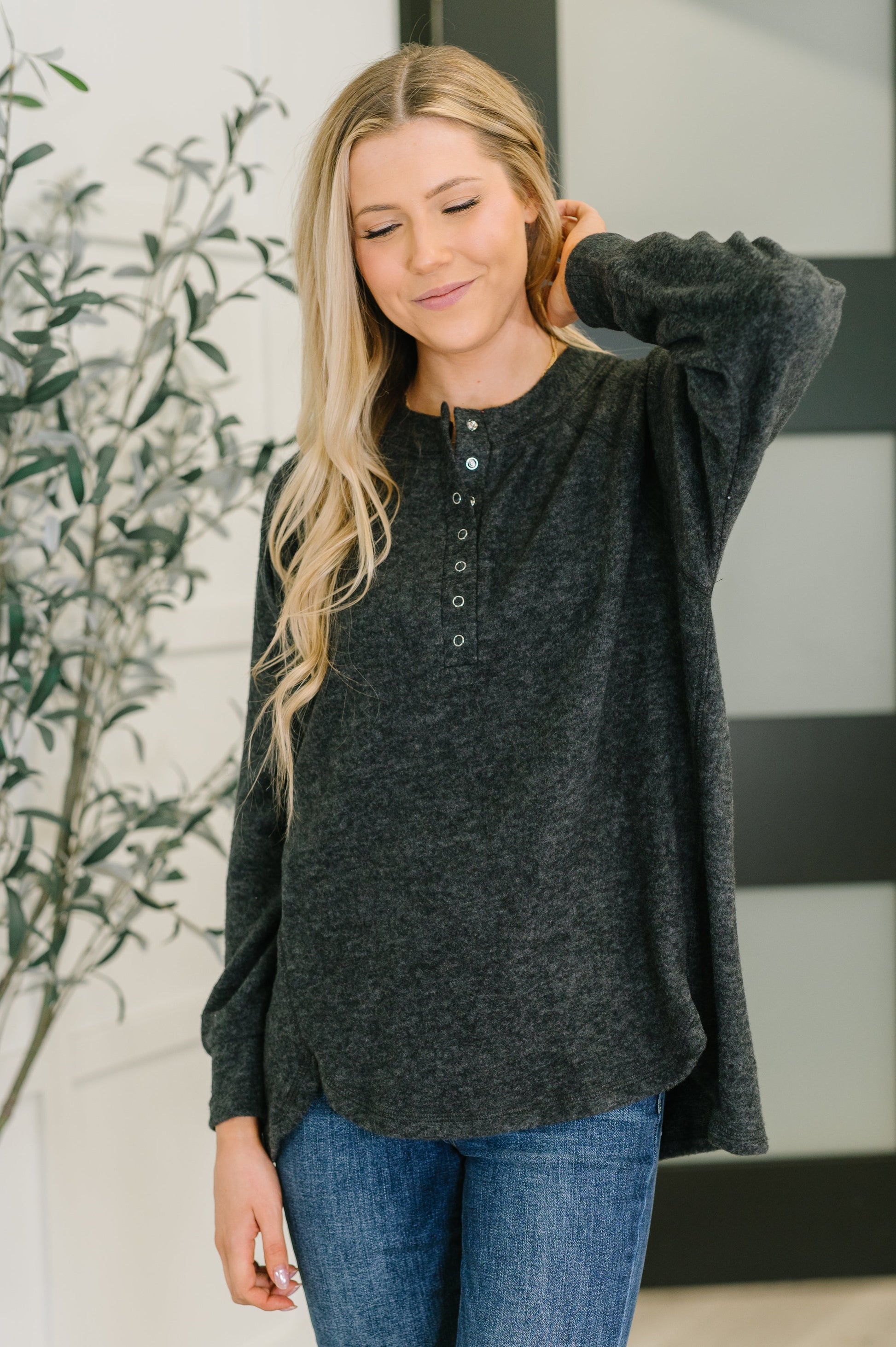 Woman wearing a black long-sleeve shirt and blue jeans, standing indoors with a plant in the background.