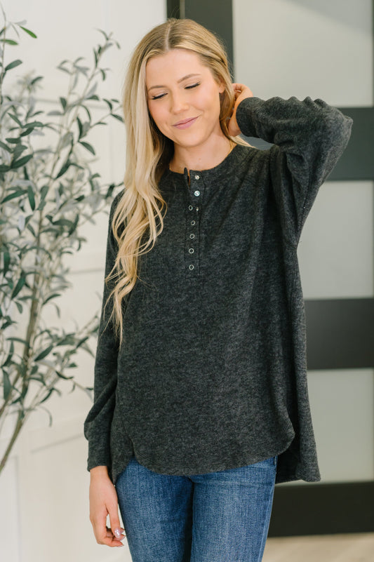 Woman wearing a black long-sleeve shirt and blue jeans, standing indoors with a plant in the background.
