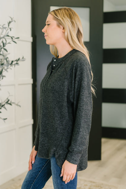 Woman wearing a black hi lo henley long-sleeve top and blue jeans standing against a black and white striped wall.