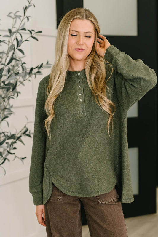 Woman wearing an olive green henley long-sleeve shirt with a blurred indoor background