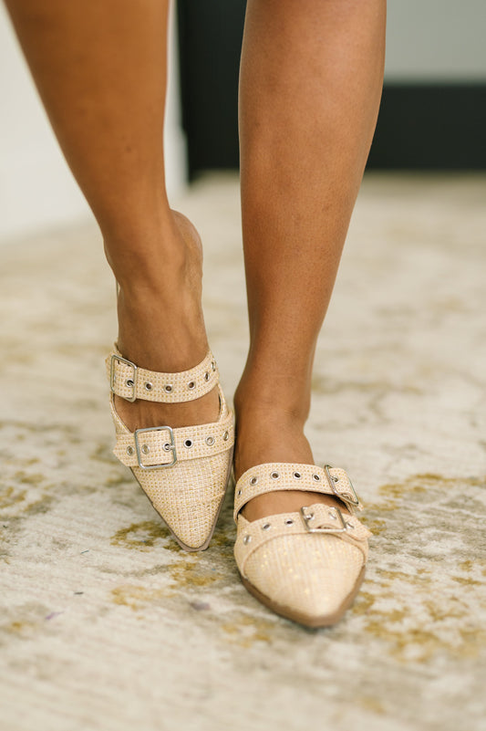 Beige pointed-toe shoes with buckles worn by a person on a textured floor.