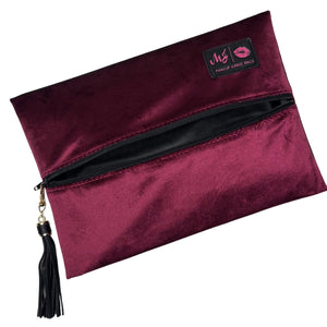 A burgundy velvet makeup bag with a black zipper, a black tassel charm, and a black brand patch on the top right.