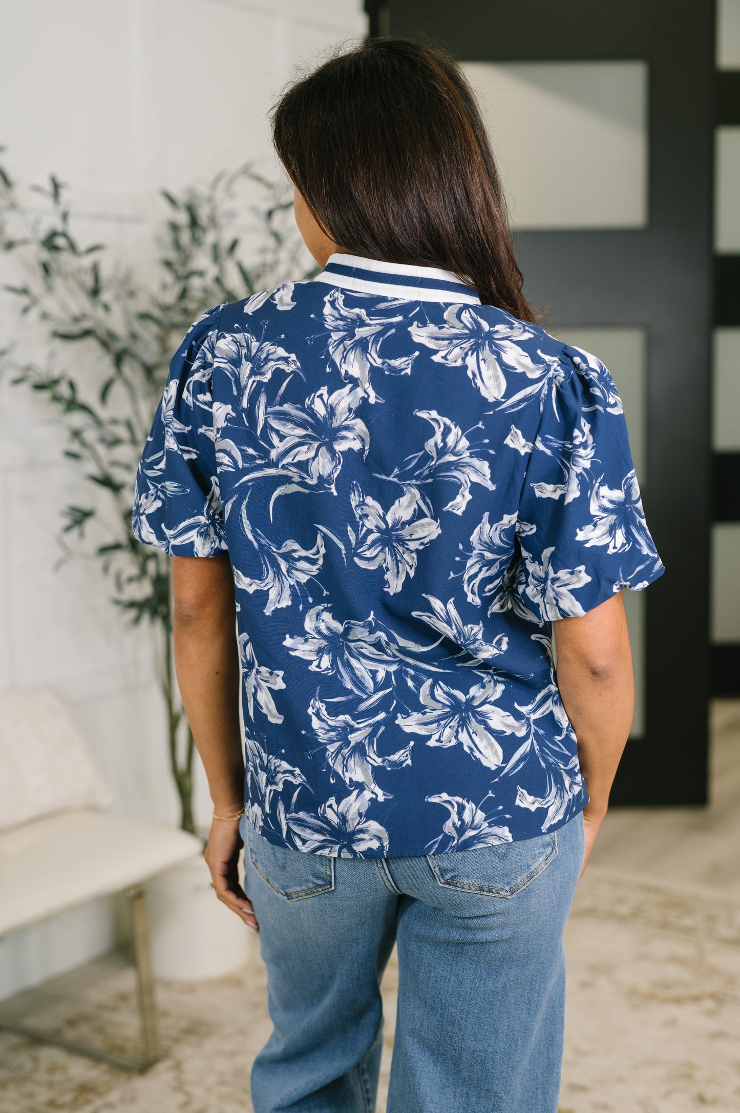 A woman viewed from behind wearing a blue short-sleeve button-up shirt with a white floral print, paired with light blue jeans in an indoor setting.