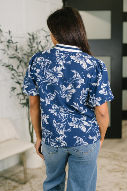 A woman viewed from behind wearing a blue short-sleeve button-up shirt with a white floral print, paired with light blue jeans in an indoor setting.