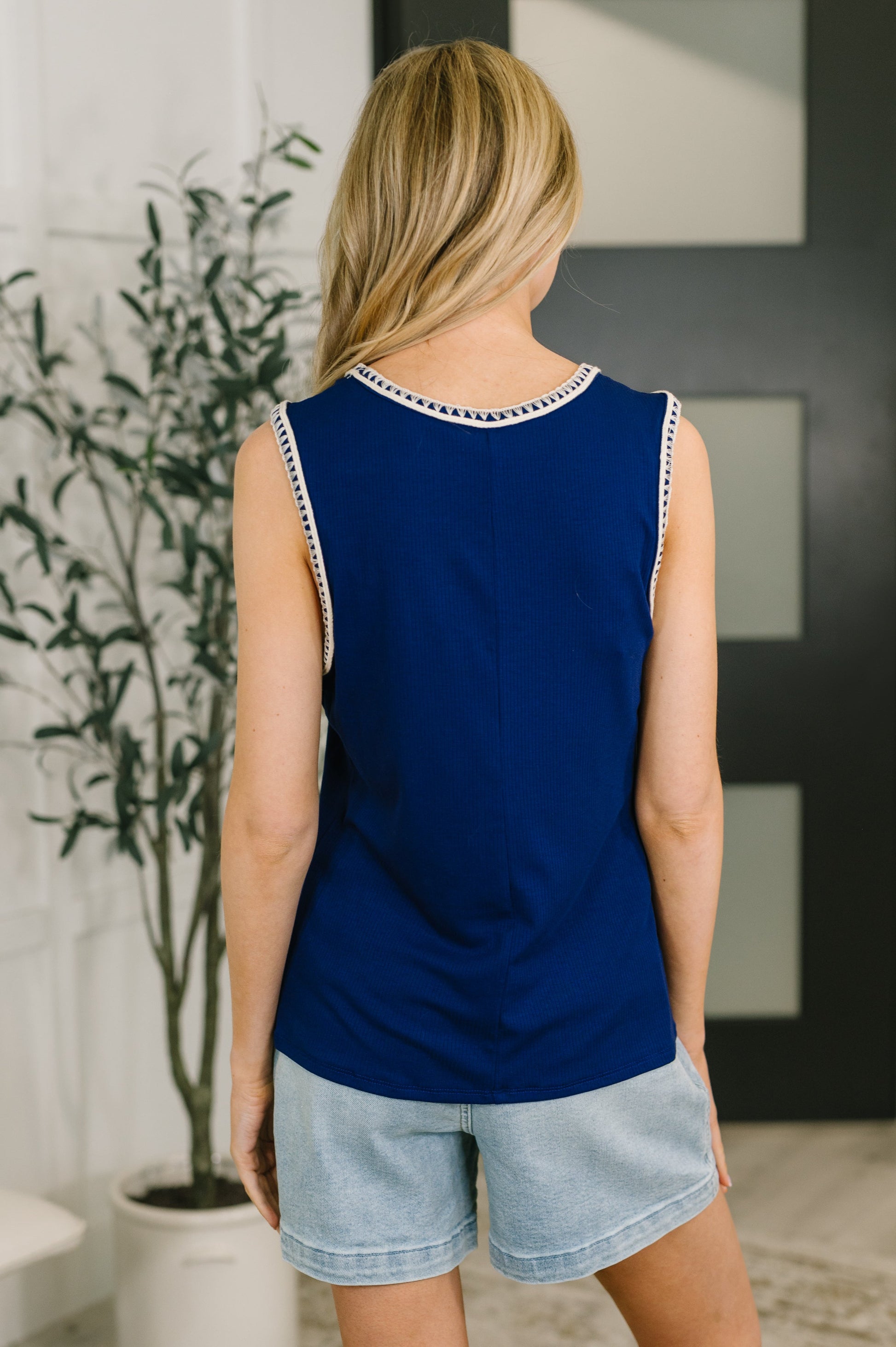 Woman wearing a blue sleeveless top with white trim and denim shorts indoors.