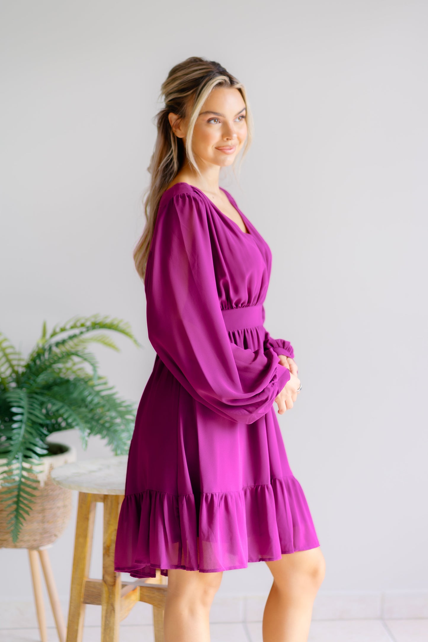 Woman wearing a purple dress standing in a room with a plant and stool.