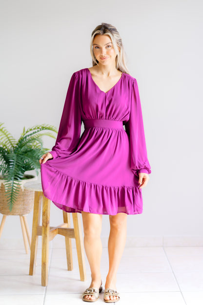 Woman wearing a purple dress standing in a room with a plant and wooden stool.