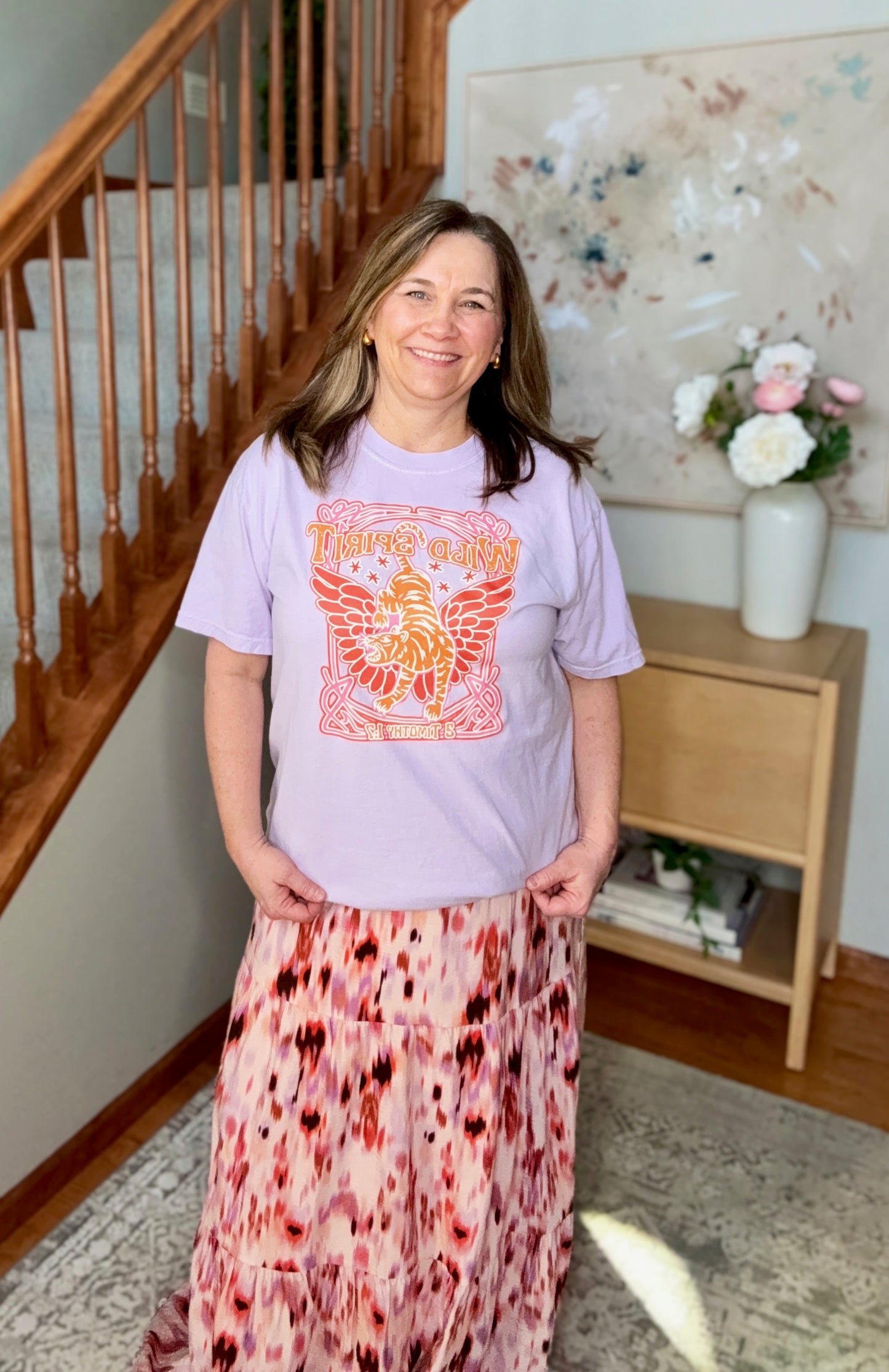 Woman wearing a light purple t-shirt with a graphic design and a patterned skirt standing in a home setting.