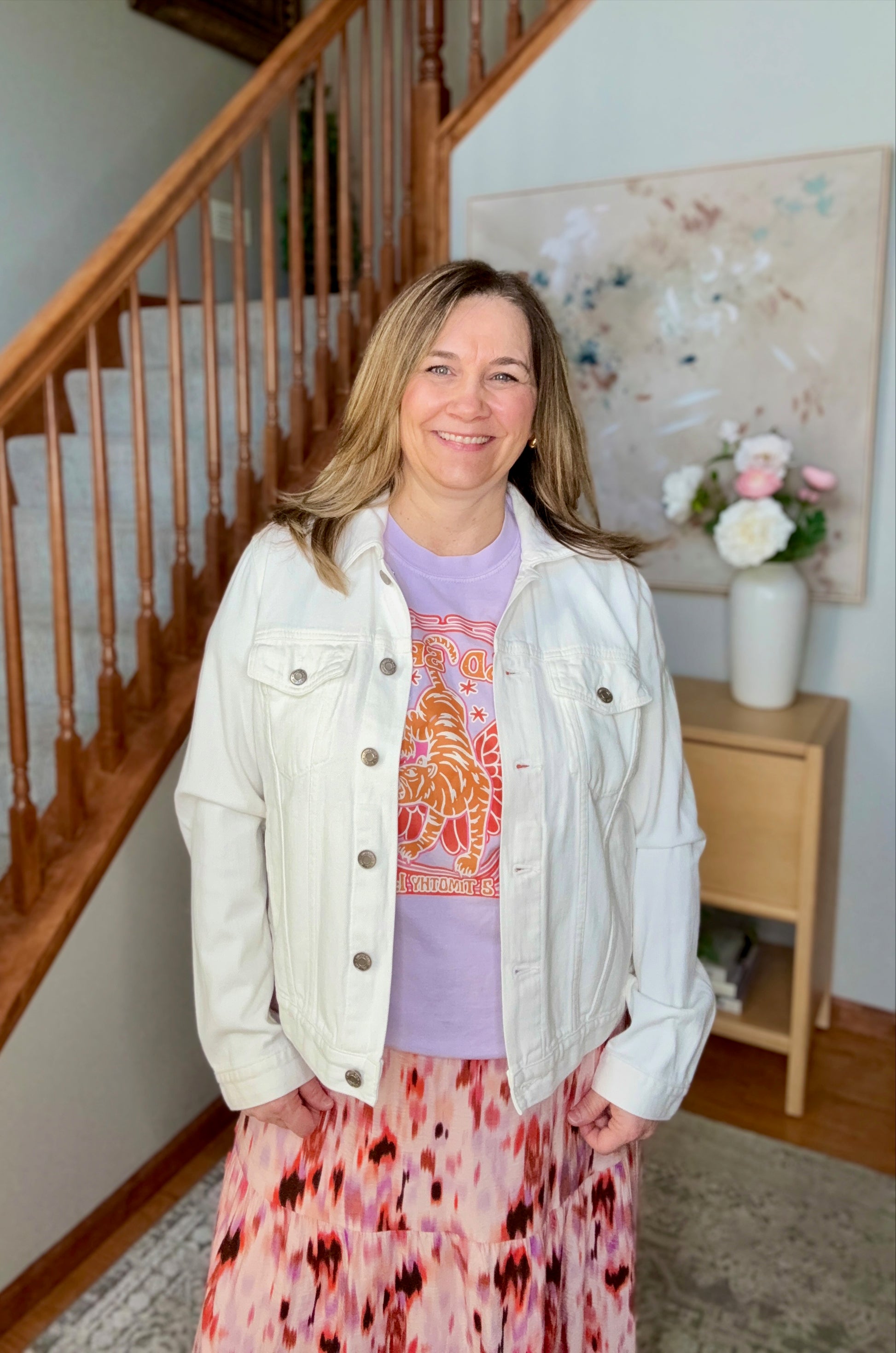 Woman standing in a home setting with a staircase and floral decorations.