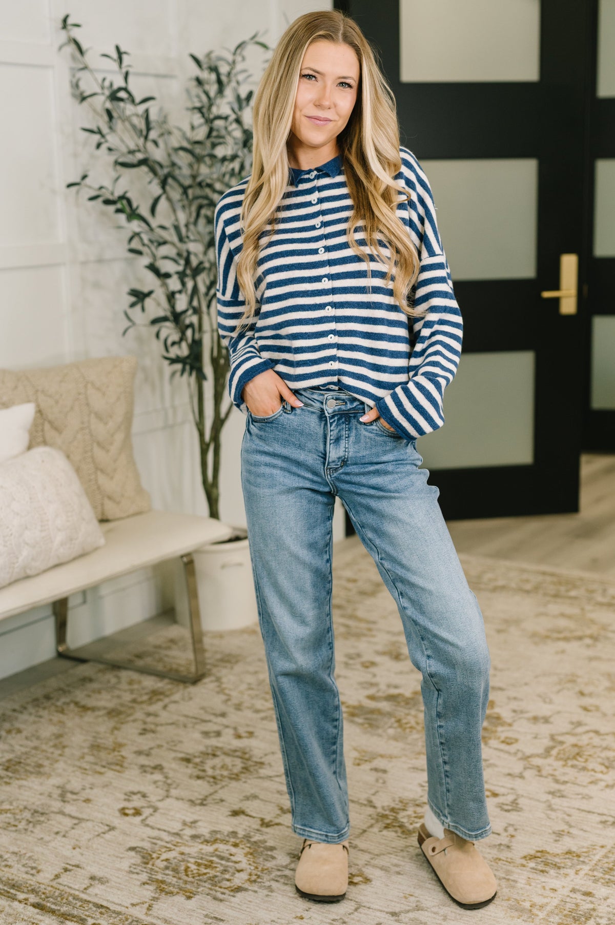 A woman stands indoors wearing a navy and white horizontally striped long-sleeve henley shirt, light-wash blue jeans, and tan slip-on shoes. The setting includes a patterned rug, a bench with pillows, and a potted plant.