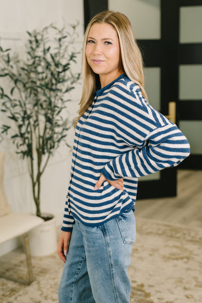 A woman modeling a blue and white horizontally striped long-sleeve knit top with light-wash blue jeans, standing in a modern indoor setting with a potted plant and a black-framed window in the background.