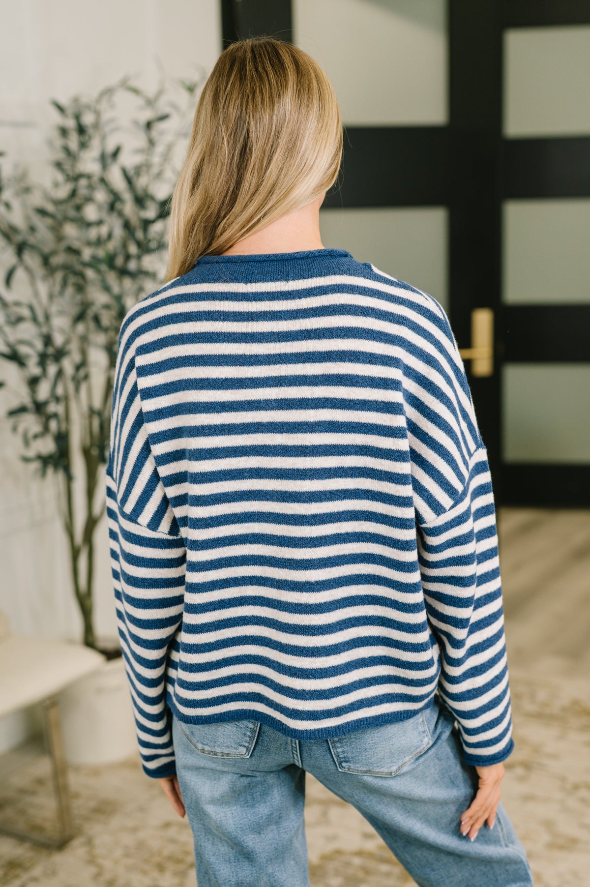 A woman viewed from behind wearing a blue and white striped knit sweater and light blue jeans in an indoor setting.