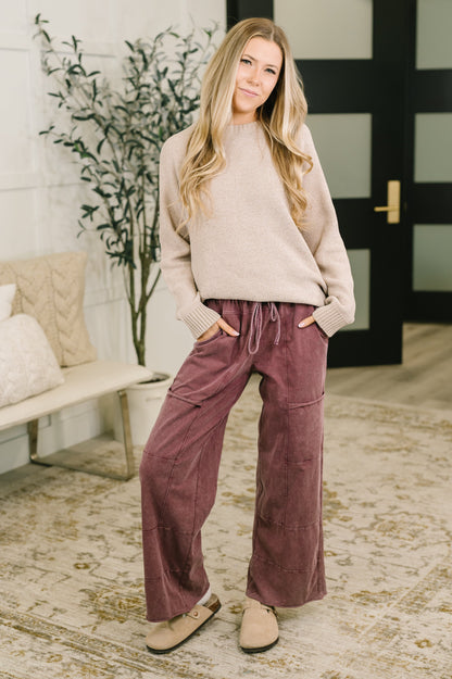A woman stands indoors wearing a beige knit sweater and dusty rose wide-leg corduroy pants with a drawstring waist, paired with tan slip-on shoes. The room has a bench, pillows, a potted plant, and a patterned rug.