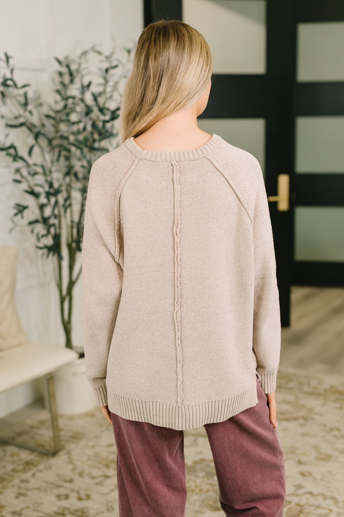 A woman viewed from behind wearing a light beige knit sweater with a central cable detail and mauve-colored pants in an indoor setting.