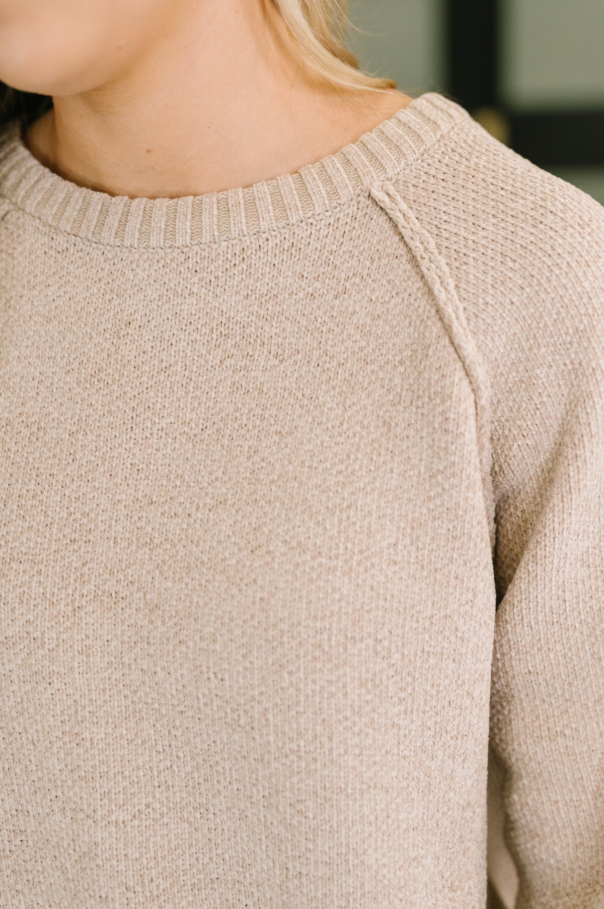 Close-up of a person wearing a light beige knit sweater, showing the ribbed crew neck, textured knit fabric, and shoulder seam.
