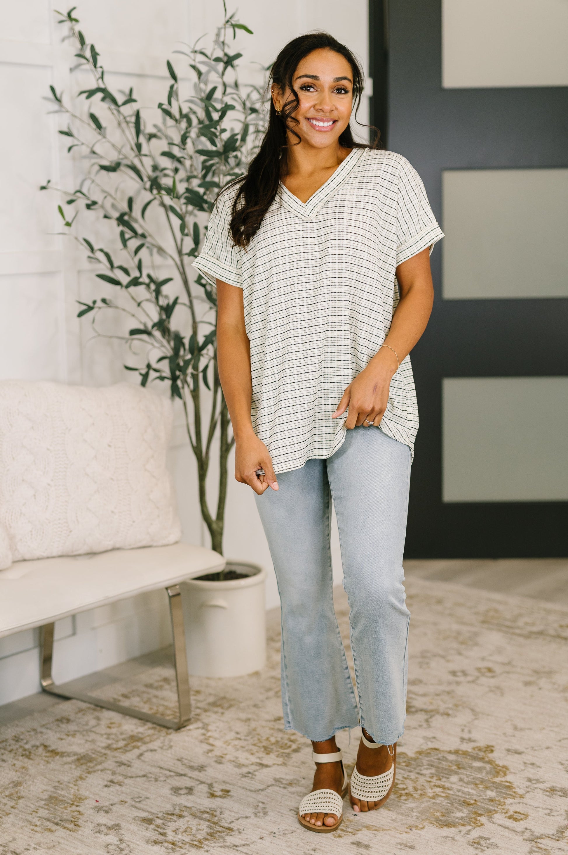 Woman wearing a patterned top and light blue jeans standing in a room with a plant and white wall.
