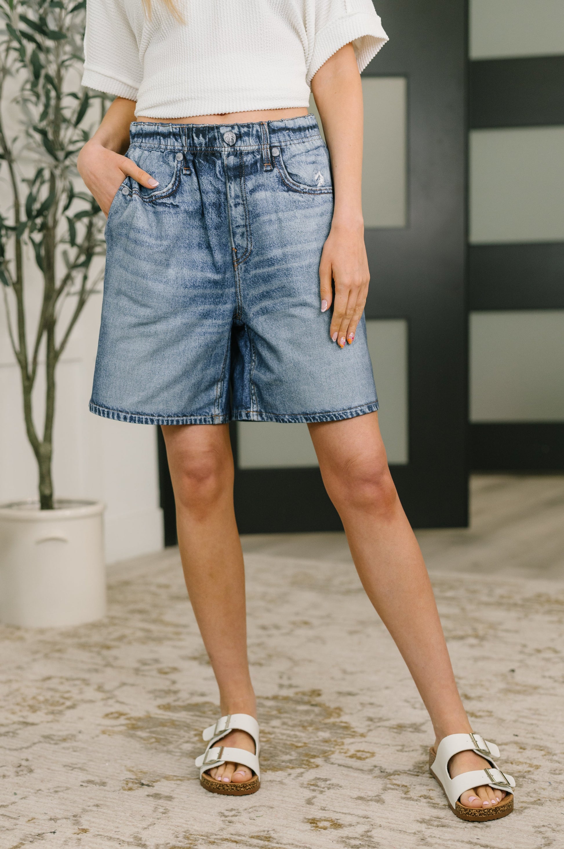 Person wearing blue denim shorts standing indoors with a plant and wall in the background