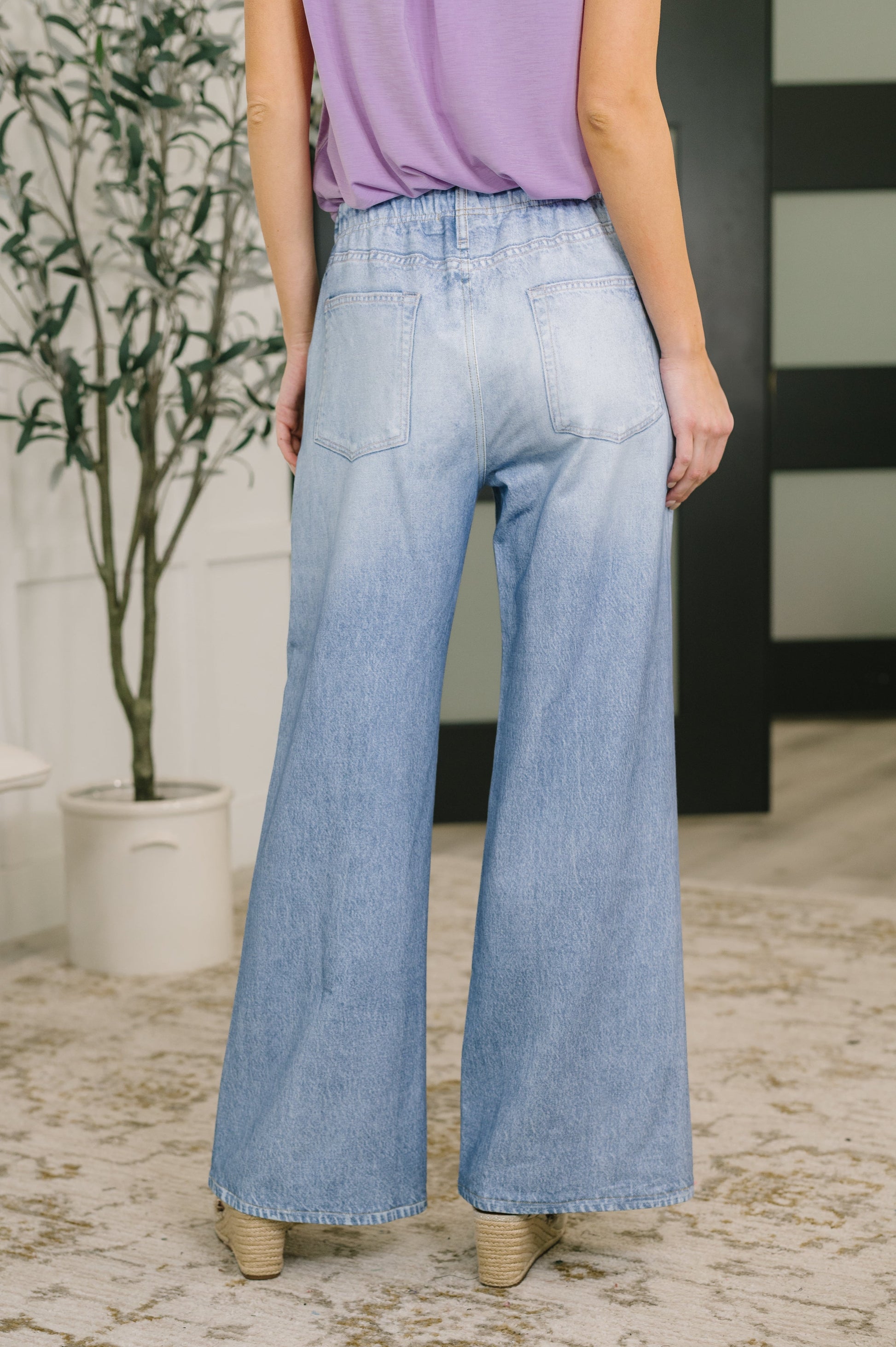 Person wearing light blue jeans and a purple top in an indoor setting with a plant and black and white striped wall.