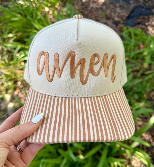 A hand holds a baseball cap outdoors with green foliage in the background. The cap has an off-white crown with metallic copper embroidered script reading 'amen' and a matching off-white and copper striped visor.