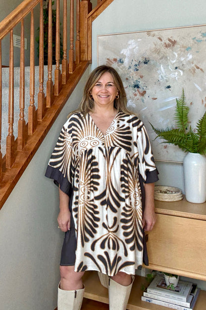 Woman in a patterned dress standing in a home interior with a staircase and decor.
