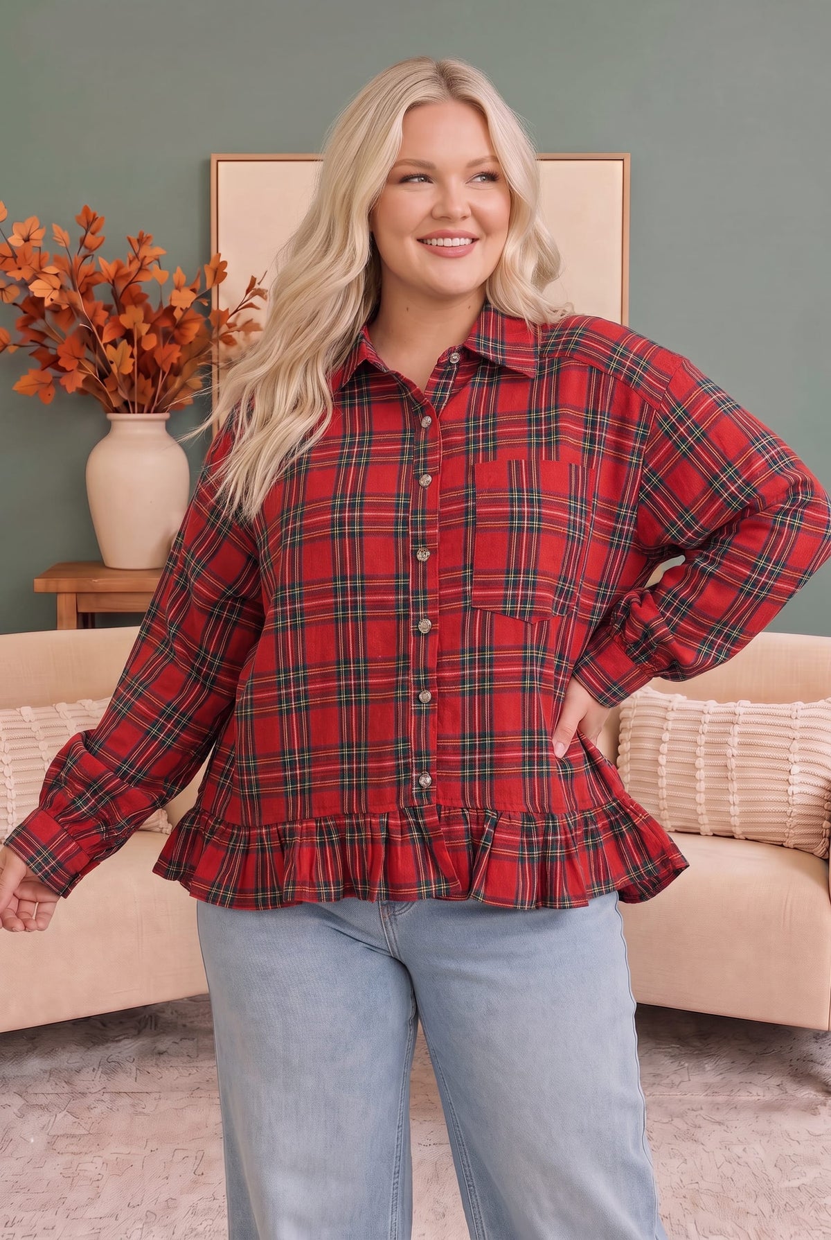 A woman modeling a red and green plaid long-sleeve button-up shirt with a ruffled hem, paired with light blue jeans, in a cozy living room setting.