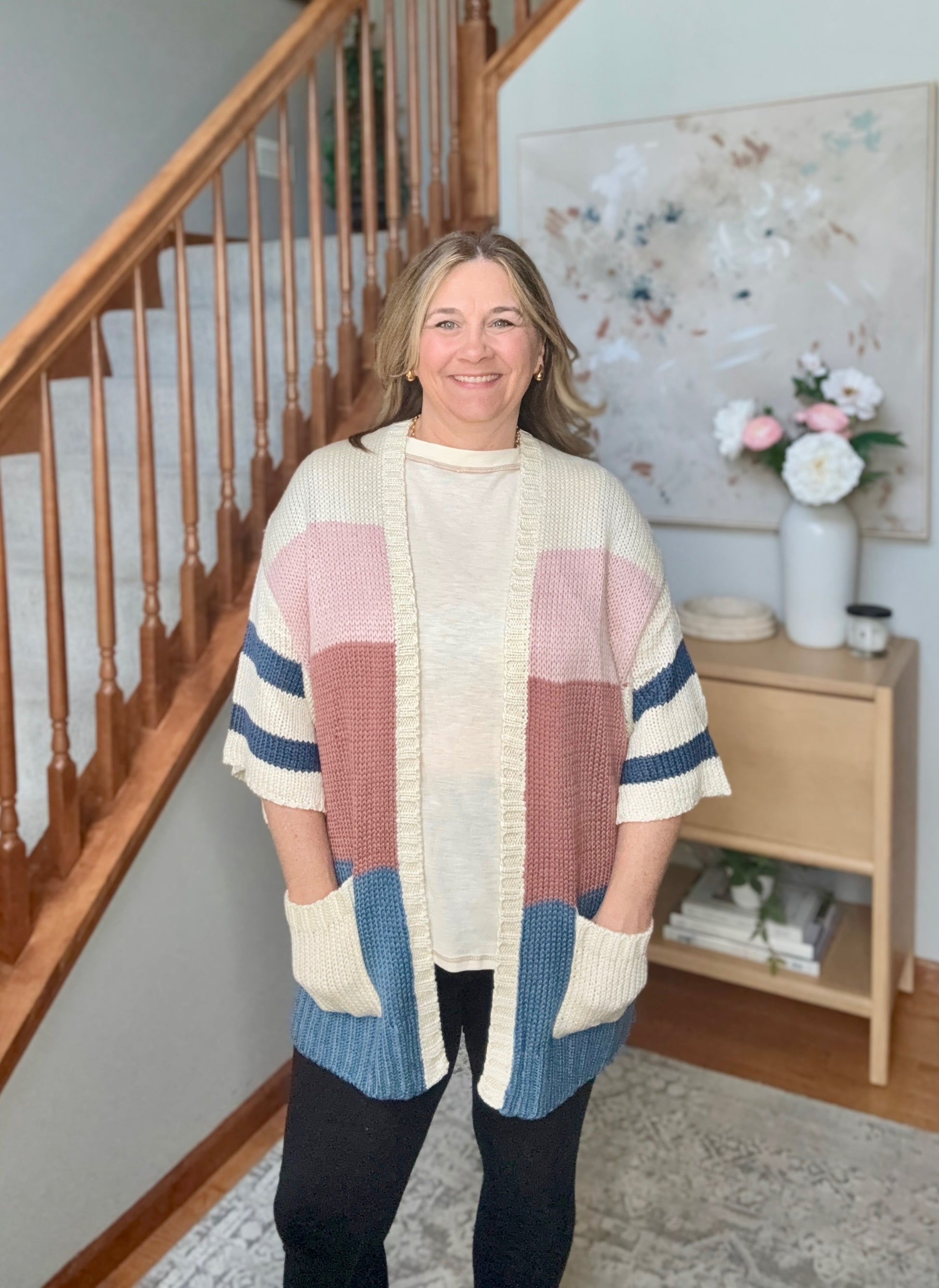 A woman wearing a chunky knit color-block cardigan with cream, pink, dusty rose, and blue panels, standing in a home interior with a staircase and side table in the background.